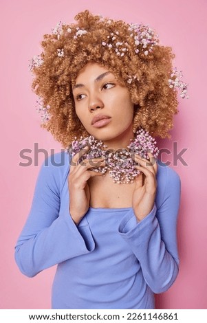 Vertical shot of thoughtful curly haired young European woman keeps hands on wreath made of dried flowers wears purple jumper focused aside isolated over pink studio background. Floral concept.