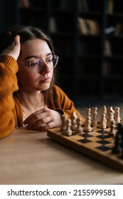 Vertical Shot Of Thoughtful Young Woman In Elegant Eyeglasses Thinking About Chess Move Sitting In Dark Library Room, Selective Focus. Pretty Intelligent Lady Playing Logical Board Game Alone At Home.