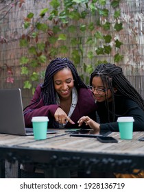 Vertical shot of two African American women sitting on a terrace outside, drinking coffee while working on various electronic devices. Laptop, tablet, smartphone. Teamwork concept.