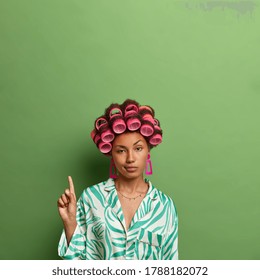 Vertical Shot Of Unimpressed Dark Skinned Woman Wears Hair Curlers And Indicates On Copy Space, Shows Blank Space Against Green Background For Your Promo, Cares About Appearance, Prepares For Date