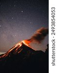 A vertical shot of the volcanic mountain Feugo erupting  at night by a starry sky in the background