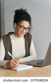 Vertical shot of young adult African American mixed race student in glasses watching online webinar on pc writing notes on desk at home modern home office. Remote distant e learning work and study.