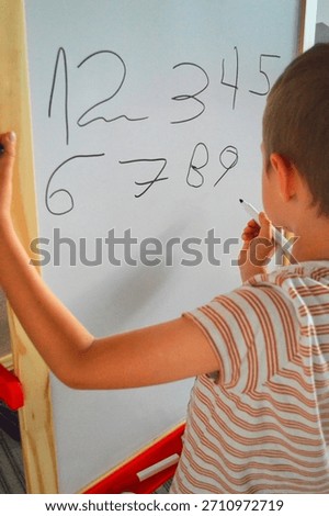 Vertical shot of a young child focused on practicing penmanship by writing the numbers 1 through 9 in black marker across a white dry-erase board. The image highlights early math education, concentration, and childhood learning activities