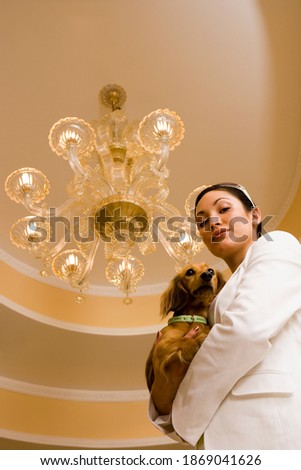 Vertical shot of a young glamorous woman carrying a dog and standing below the chandelier.