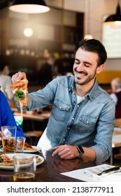 Vertical Shot Young Hipster Caucasian Man Sitting At Table In Restaurant Laughing Because His Piece Of Pizza Has Stringy Cheese.