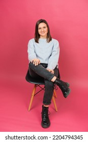 Vertical Shot Of A Young Smiling Woman Is Sitting On A Chair Holding One Leg Up In A Pink Studio.