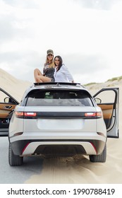 A Vertical Shot Of Young Spanish Girls Having Fun While Sitting On The Top Of Their Car On The Beach