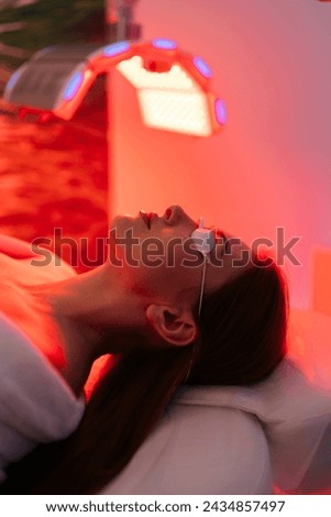 Vertical shot of young woman having red LED light facial photodynamic therapy treatment in beauty salon. Luxury medical clinic using phototherapy device with red light wavelength for acne treatments.