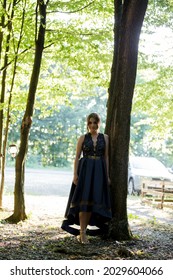 A Vertical Shot Of A Young Woman Standing In A Tree Shade In A Blue Dress