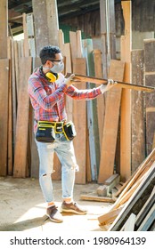 Vertical Side View Of Caucasian Male Carpenter In Safety Goggles, Ear Muffs On His Neck, Wearing Face Mask Standing Checking Surface And Quality Of A Wooden Plank That Held In Hands In A Wood Factory.