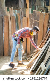 Vertical Side View Of Caucasian Male Carpenter In Safety Goggles, Ear Muffs On His Neck, Wearing Face Mask Bending Over Holding A Wooden Plank From The Pile In Front Of Him In A Wood Factory.