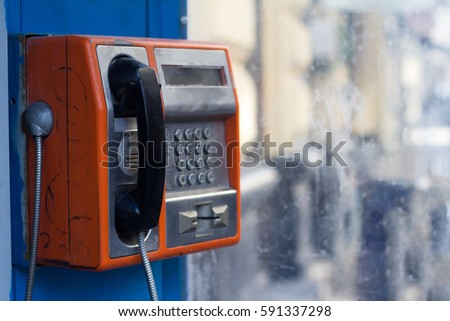 Vertical side view of damaged orange public phone with near a dirty window with selective focus