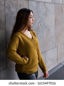 Vertical Side View Of Pretty Brunette Woman With Waist Length Hair In Mustard Coloured Sweater Standing In Front Of Stone Wall With One Hand On Hip And Determined Expression