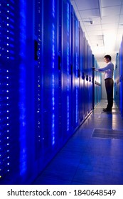 A Vertical Side View Shot Of An IT Technician Working And Checking System In The Aisle Of A Data Center Room With A Row Of Glowing Network Server Cabinets On One Side