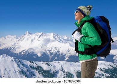 A Vertical side view of a woman smiling at the sun wearing a backpack and heavy winter wear standing on a background of a snow mountain