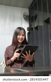 A vertical, slightly low-angle shot of a young Asian woman looking down intently at a black tablet device with a soft smile. She holds a dark iced drink in her left hand. The modern background features a mix of industrial elements, including a corrugated wall and a wall-mounted fan. Perfect for e-learning, mobile browsing, or modern lifestyle imagery.