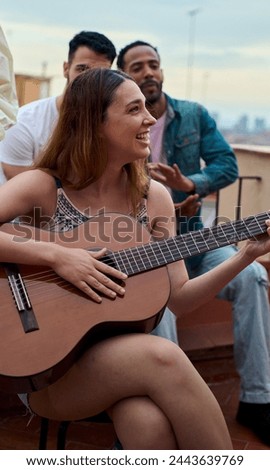 Vertical. Smiling Caucasian blonde woman happy plays flamenco guitar while her group multiracial friends clap at rooftop party. Young people gathered together enjoying summer vacations