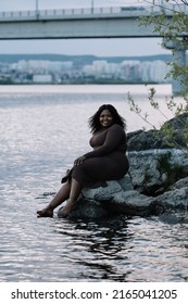 Vertical smiling playful barefoot curly plump full-figured Afro American interracial woman sit by the sea or river coast, wash legs and rest. Mixed race ethnicity, diversity. Windy summer weekends