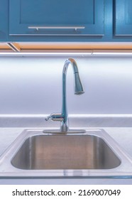 Vertical Stainless Sink With Single Faucet And Marble Counter. Kitchen Sink Against The Marble Backsplash With Lights Below The Gray Wall Cabinets Above.