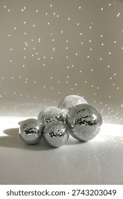 Vertical studio shot of a cluster of silver mirror disco balls of various sizes resting on a white surface. Bright sunlight hits the mosaic glass, creating a magical pattern of scattered glowing light reflections on the wall behind them. Perfect for party invitations, New Year's Eve celebrations, and retro music concepts