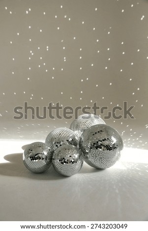 Vertical studio shot of a cluster of silver mirror disco balls of various sizes resting on a white surface. Bright sunlight hits the mosaic glass, creating a magical pattern of scattered glowing light reflections on the wall behind them. Perfect for party invitations, New Year's Eve celebrations, and retro music concepts