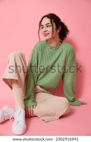 Vertical studio shot of teenage girl sitting on floor in front of a pink background wearing green sweater, beige pants, white sneakers and looking away has positive expression smiling and posing.