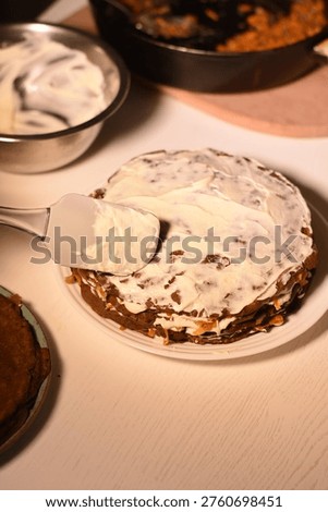 Vertical top angle view of spreading mayonnaise on a layer of liver cake with spatula, bowls with liver pancakes and mayonnaise on light table nearby