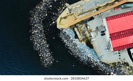 Vertical top down drone view of a waterfront pier with red roof and curved rock breakwater in Naples, Italy.