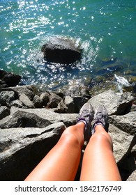 Vertical And Top Down Shot Of A Pair Of Legs And Feet In Sneakers (tennis Shoes), On Rocks Above A Lak, On A Sunny Day