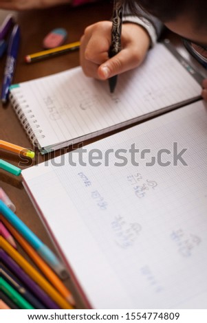 Vertical top view shot of Kid hands doing math homework on a wooden table with color crayons, pencils and notebooks by a white child faceless unrecognizable wearing glasses. Study at home concept.