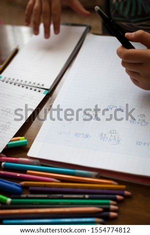 Vertical top view shot of Kid hands doing math homework on a wooden table with color crayons, pencils and notebooks by a white child faceless unrecognizable wearing glasses. Study at home concept.