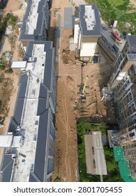 Vertical top-down aerial view of an active earthwork zone at a residential construction site in Zhengzhou, China. Excavators, a truck crane and heavy machinery operate on bare dirt ground with pipe trenches and material piles. Finished apartment blocks stand on the left, scaffolded unfinished buildings on the right.