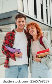 Vertical. Two cheerful classmates posing together outdoors, smiling warmly at the camera while holding books and backpacks, showcasing a friendly and happy student life outside their university campus
