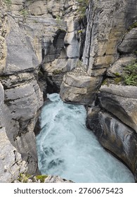A vertical view captures a raging river slicing through a rugged, rocky landscape as it carves fresh paths toward Banff National Park. Towering cliff sides—etched with years of erosion from the same relentless water—reveal a dramatic chasm, offering a striking glimpse into the raw geology of Alberta.