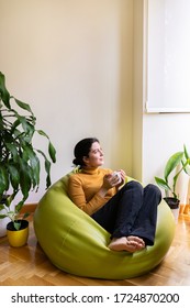 Vertical view of casual woman at home drinking herbal tea sit on a green pouf. Quarantine home lifestyle. Isolated woman drinking a hot beverage indoors. Healthy lifestyle. Spring and autumn seasonal.