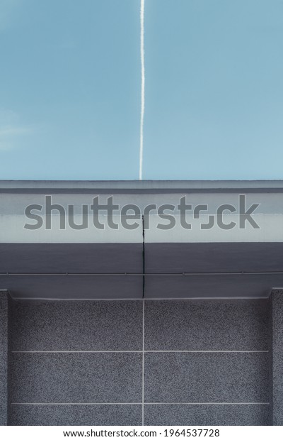 Vertical view of a concrete wall of a building with\
lines dividing cement facade and roof blocks, and a vertical line\
which continues with a real airplane contrail on the blue sky going\
directly up