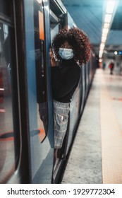 Vertical View Of A Curly Young Black Female In A Virus Protective Facial Mask, She Is Hanging Out Of The Door Of A Metro Train On A Modern Subway Station; Shallow Depth Of Field, Selective Focus