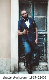 Vertical view of a dapper bald mature black guy with a beard, in a suit with a bow tie and leather bag, leaning against the wall on the stoop of an antique building after leaving the wooden door