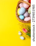 Vertical view of Easter wicker basket full of colorful eggs and spring flowers on bright yellow background, perfect for spring celebrations and greetings.