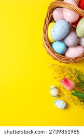 Vertical view of Easter wicker basket full of colorful eggs and spring flowers on bright yellow background, perfect for spring celebrations and greetings.
