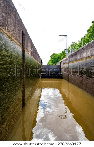 Vertical view inside a canal lock chamber on a waterway near Bamberg in Bavaria, Germany, showing high concrete walls with algae growth and brown water reflecting the cloudy sky in daylight. Trees and a street lamp are visible above the lock structure, emphasizing the industrial navigation infrastructure in an urban environment.