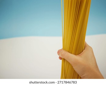 Vertical view of a large bundle of uncooked spaghetti pasta held by a hand on blue and white background, modern food photo of dry Italian ingredients This food photo set shows uncooked spaghetti pasta on a blue and white background. Some images feature a hand holding a large bundle of long dry pasta, others display the spaghetti laid flat on a white surface. Ideal for Italian cuisine concepts, cooking ingredients, diet articles and food design.