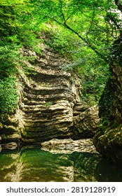 A vertical view of layered limestone cliffs and an emerald pool in a Georgian forest gorge conveys a wild, peaceful and secluded summer atmosphere