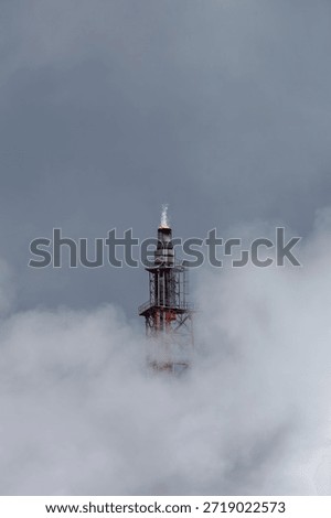 Vertical view of lone flare stack emitting flame with sharp white plume. Surrounded by wafting vapor clouds set to overcast gray-blue sky background, scene portrays environmental impact of industry.