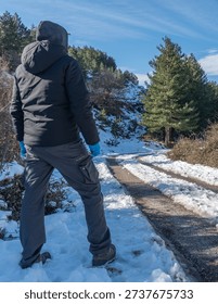 Vertical view of a man seen from behind standing on the edge of a snow covered mountain road. Blue sky with few clouds above a winter mountain landscape.
