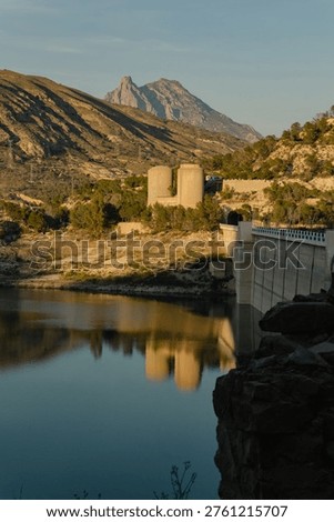 A vertical view of the massive concrete wall and green spillway gates of the Amadorio Dam in Alicante. The still reservoir water reflects the industrial scale of this civil engineering project. Arid mountains are visible above the structure. This facility is critical for water management and irrigation in the Valencian Community of Spain.