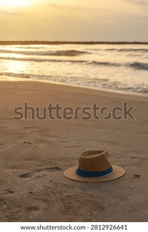 Vertical view of a Panama-style hat resting on golden sand beside a human footprint, with the calm sea in the background on a warm summer morning, highlighting peaceful beach landscapes, coastal tourism, and relaxing holiday destinations