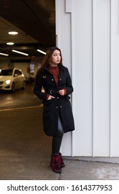 Vertical View Of Pretty Brunette Woman With Waist Length Hair Wearing Black Coat And Leather Pants Standing Waiting At The Entrance Of An Indoor Parking Building