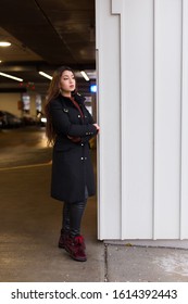 Vertical View Of Pretty Brunette Woman With Waist Length Hair Wearing Black Coat And Leather Pants Standing With Arms Crossed Waiting At The Entrance Of An Indoor Parking Building