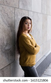 Vertical View Of Pretty Brunette Woman With Waist Length Hair In Mustard Coloured Sweater Standing In Front Of Stone Wall With Arms Crossed While Staring With Stern Expression